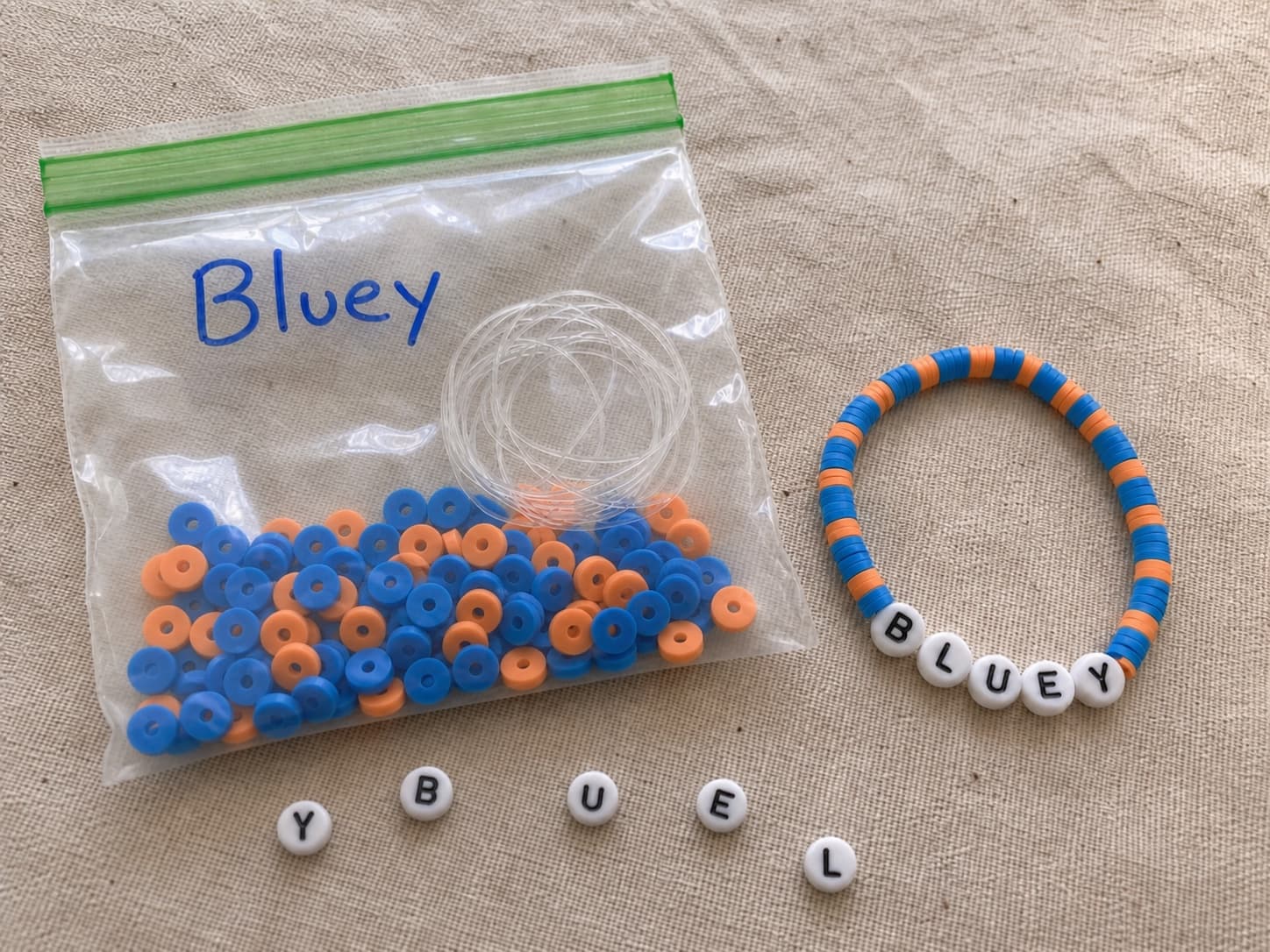 Handmade blue and orange BLUEY bracelet beside its clear DIY bead kit and loose letter beads