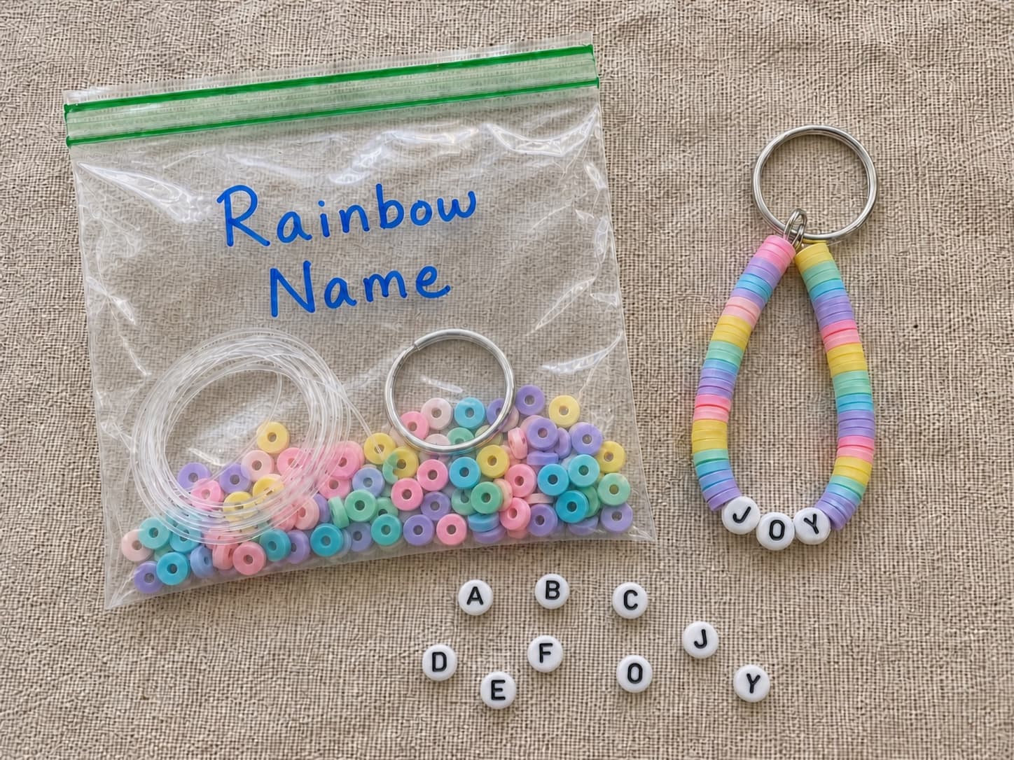 Finished rainbow JOY keychain beside its clear DIY bead kit and loose alphabet beads