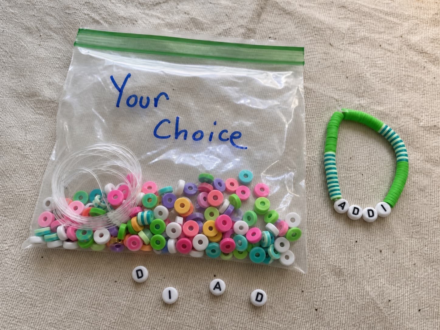 Handmade green and teal ADDI bracelet beside its clear DIY bead kit and loose letter beads